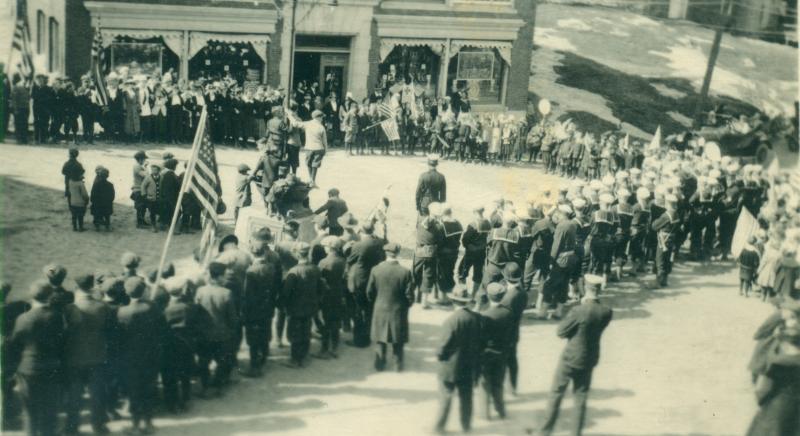 The historical society has dozens of parade photos, almost all undated and showing Townsend Avenue businesses hung with bunting. This view may have been the parade gathering on the first Armistice Day. The impromptu parade(s) that sprang up that morning upon the news of peace would not have allowed time to hang bunting. Boys have climbed on the Townsend Avenue horse trough in Bank Square in front of the bank for a better vantage. The Coast Patrol men are all in uniform, and the coats show the weather is cold. Part of the area where the Kaiser bonfire was located, the ledges between the bank and the brick house, are visible.