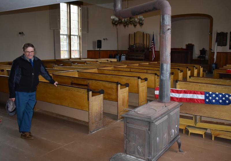 Selectman Allen Greene inside Woolwich’s Nequasset Meeting House. At right is one of three woodstoves once used to heat the building. Beneath the pew are footstools. PHIL DI VECE/Wiscasset Newspaper
