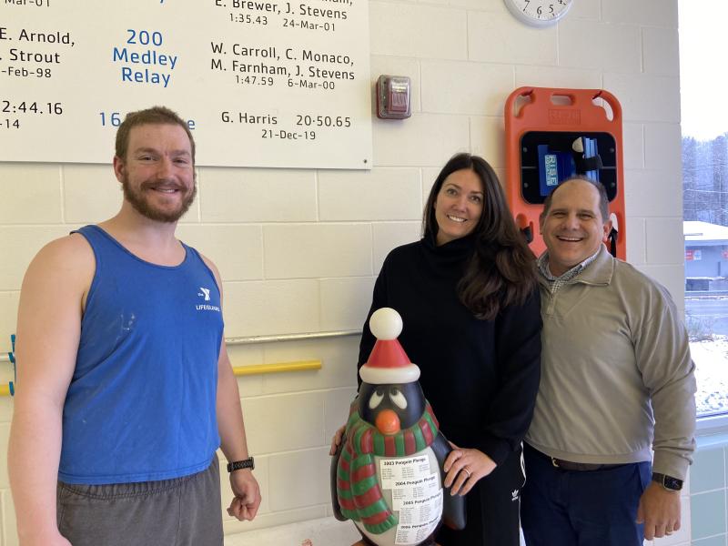 Y Aquatics Director David Washburn, left, stands beside the Penguin Plunge mascot and this year’s Penguin Plunge Grand Marshals, Medea and Bruce Harris. Note their son Graham’s 2019 swim record on the wall behind them! Courtesy of BRYMCA