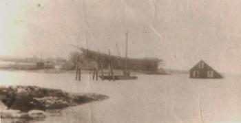 This April 1920 view shows the Machon house bobbing its way out of Mill Cove on its unmanned voyage to Tumbler Island. Small scows, such as that by the pilings, were fairly common. Perhaps it's the leaking one that set the house free, though it looks too small for half a house. The vessel being built on the shore of McFarlands Point at the Atlantic Coast Company is the 4-masted schooner James W. Howard, launched July 5, 1920. Courtesy of Lucille Machon