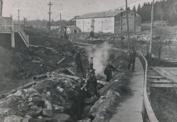 By late November 1912 Elbridge Giles's waterline pipe-laying crew had rounded Mill Cove and was headed for West Harbor. The photo's largest feature is the Mill Cove Ice Company's icehouse, with its elevator rising to the top. The site's gentle hill was blasted level and covered with condominiums in the late 1980s. The 1900s ice run to the deepwater wharf is apparent; it looks like a little sidewalk running outside the obvious sidewalk. The poles in the cove constitute the fish weir of Clarence or Charles Hodgdon, meant to trap herring and alewives. Courtesy of Barbara Rumsey
