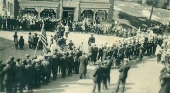 The historical society has dozens of parade photos, almost all undated and showing Townsend Avenue businesses hung with bunting. This view may have been the parade gathering on the first Armistice Day. The impromptu parade(s) that sprang up that morning upon the news of peace would not have allowed time to hang bunting. Boys have climbed on the Townsend Avenue horse trough in Bank Square in front of the bank for a better vantage. The Coast Patrol men are all in uniform, and the coats show the weather is cold. Part of the area where the Kaiser bonfire was located, the ledges between the bank and the brick house, are visible.
