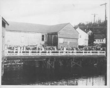 In 1951 the lumper crew and anyone else they could find in both yards (about 35 men) carry a mast across the East Boothbay bridge from the Hodgdon yard to Goudy's. The bridge is now a culvert; the old Hodgdon tidemill, at the inlet/outlet of the mill pond, is behind the men. It was torn down in 1959. Courtesy of the Boothbay Region Historical Society