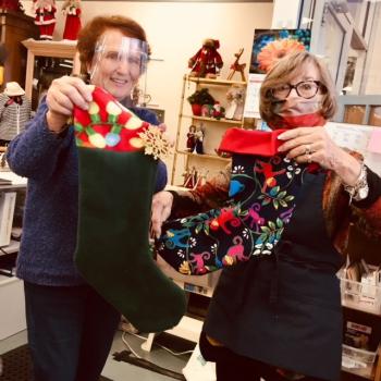 Linda Clapp and Laura Honey (right,) hold examples of the lovely stockings prepared for the residents of the Village with the colorfully stocked Thrift Shop in the background. Courtesy of the St Andrews Auxiliary
