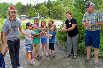 Kids gather around a pair of clippers to help Executive Director Nick Ullo, right, and Environmental Educator Tracey Hall cut the branch and officially open the playground. At left is John Copeland, owner of Kennebec Trail Company which did all the construction of the playground. Peter Southwick photo