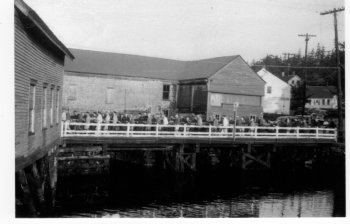 More than 50 men carry a mast south across the bridge in East Boothbay in 1951, and prepare to make the turn into Goudy & Stevens shipyard. In the background is the old Hodgdon tidemill perched on the entrance to the mill pond from the Damariscotta River. The tidemill was regrettably torn down to widen the road and bridge about 1959. Courtesy of Boothbay Region Historical Society