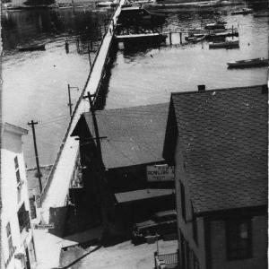 This mid-1900s shot is taken from the roof of the formerly taller and flat-roofed bank building in Bank Square. It looks east to the Romar Bowling Lanes run by Charlie Rowe, on the right, and the Blue Ship restaurant, run by his wife Kay Marr Rowe, on the left. The Romar name derives from combining Rowe and Marr. Across the footbridge, the little low building is the Bryan Rowe store. There is no good way to get a good view of the foreground buildings on their By-Way locations. Courtesy of Barbara Rumsey